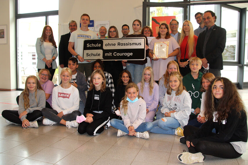 Eine Gruppe mit mehreren Kindern und Erwachsenen halten ein Schild mit der Aufschrift 'Schule ohne Rassismus - Schule mit Courage' in den Händen.