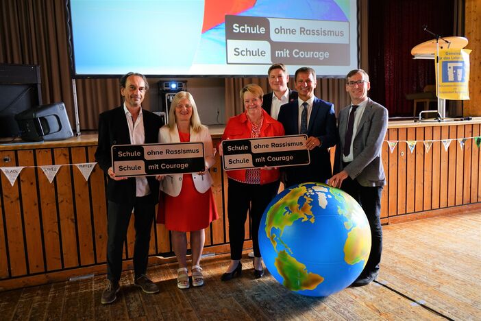6 Personen halten Logos in den Händen um Thema 'Schule ohne Rassismus - Schule mit Courage'